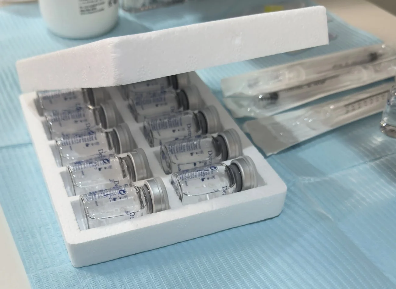 An opened polystyrene tray of sealed vials of treatment solution, with sterile packaged syringes and a further vial on a blue clinical couch cover